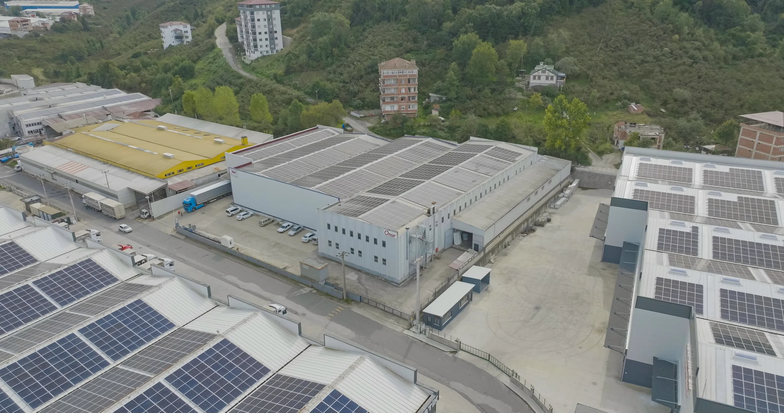 Havadan görünüm: çatıları güneş panelleriyle kaplı endüstriyel binalar, aralarında Onur logolu gri bir bina; arkada ağaçlık yamaç ve konut blokları.